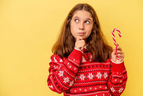 Little caucasian girl holding Christmas stick isolated on yellow background looking sideways with doubtful and skeptical expression. - Фото, изображение