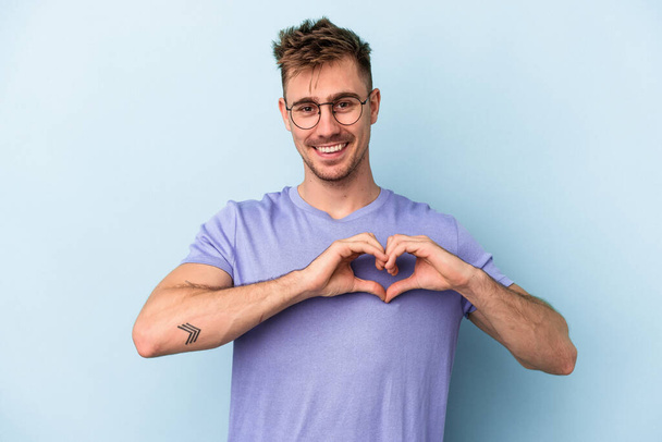 Jonge blanke man geïsoleerd op blauwe achtergrond glimlachend en toont een hartvorm met handen. - Foto, afbeelding