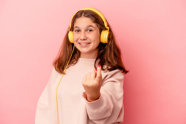 Kis kaukázusi lány zenét hallgat elszigetelt rózsaszín háttérrel mutogatva ujjal rád, mintha meghívó közelebb jönne.. - Fotó, kép
