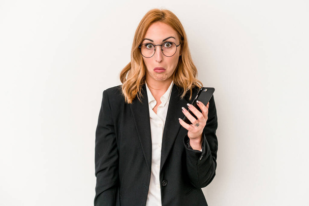 Young business caucasian woman holding mobile phone isolated on white background shrugs shoulders and open eyes confused. - Foto, afbeelding