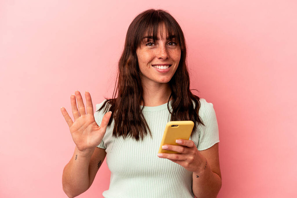 Young Argentinian woman holding a mobile phone isolated on pink background smiling cheerful showing number five with fingers. - Φωτογραφία, εικόνα