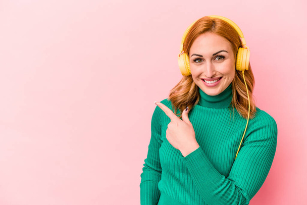 Молодая белая женщина слушает музыку, изолированную на розовом фоне улыбаясь и указывая в сторону, показывая что-то в пустом пространстве. - Фото, изображение