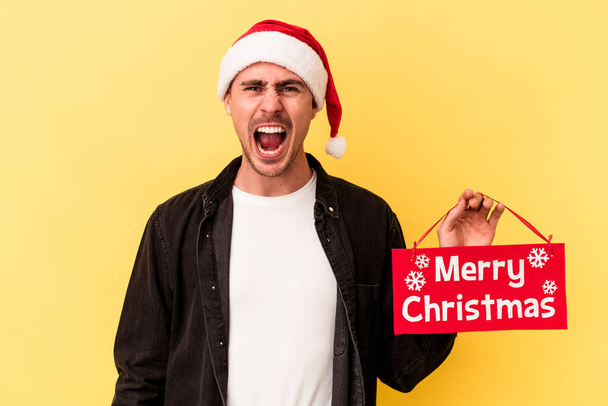 Junger kaukasischer Mann hält ein Plakat mit frohen Weihnachten auf gelbem Hintergrund und schreit sehr wütend und aggressiv. - Foto, Bild