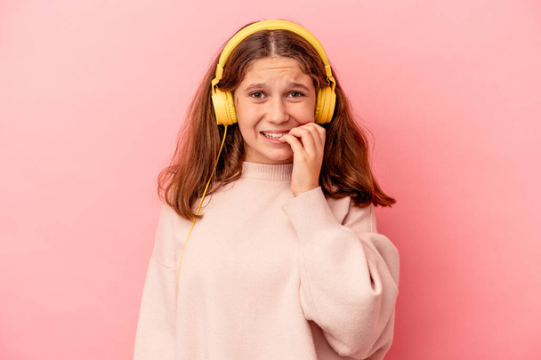 Malá běloška poslouchá hudbu izolovanou na růžovém pozadí kousání nehty, nervózní a velmi úzkostlivý. - Fotografie, Obrázek