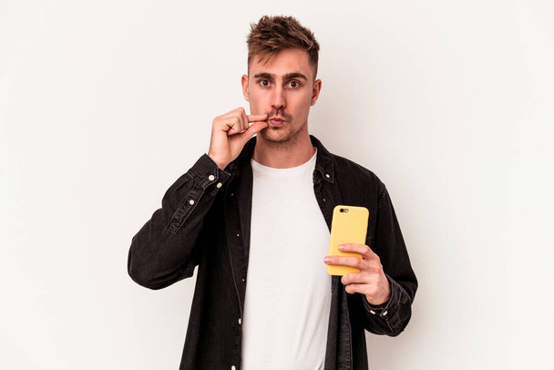 Young caucasian man holding a mobile phone isolated on white background with fingers on lips keeping a secret. - Photo, Image