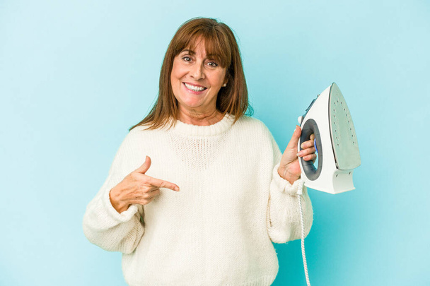 Middle age caucasian woman holding a iron isolated on blue background person pointing by hand to a shirt copy space, proud and confident - Foto, immagini