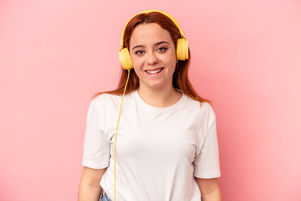 Junge kaukasische Frau hört Musik isoliert auf rosa Hintergrund glücklich, lächelnd und fröhlich. - Foto, Bild