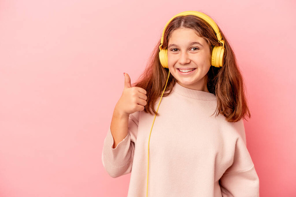 Pequeña chica caucásica escuchando música aislada sobre fondo rosa sonriendo y levantando el pulgar hacia arriba - Foto, imagen