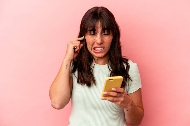 Молода аргентинка тримає мобільний телефон, ізольований на рожевому фоні, закриваючи вуха руками.. - Фото, зображення