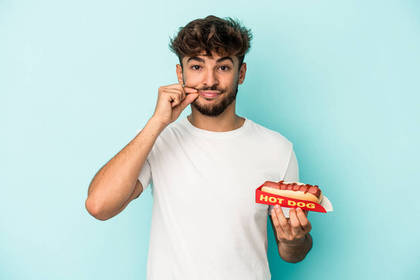 Young arab man holding a hotdog isolated on blue background with fingers on lips keeping a secret. - Photo, image