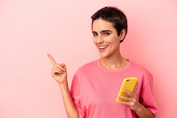 Young caucasian woman holding mobile phone isolated on pink background smiling and pointing aside, showing something at blank space. - Valokuva, kuva