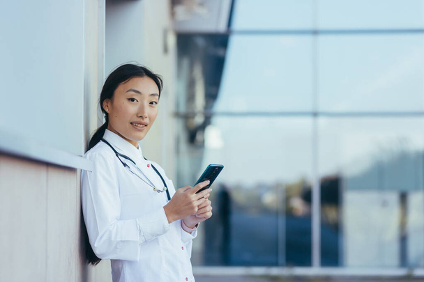 Happy female doctor, smiling and rejoicing, Asian woman uses the phone, during a break near the clinic medical apps, looking at screen, working online, chatting, consulting patient, telemedicine concept - Zdjęcie, obraz