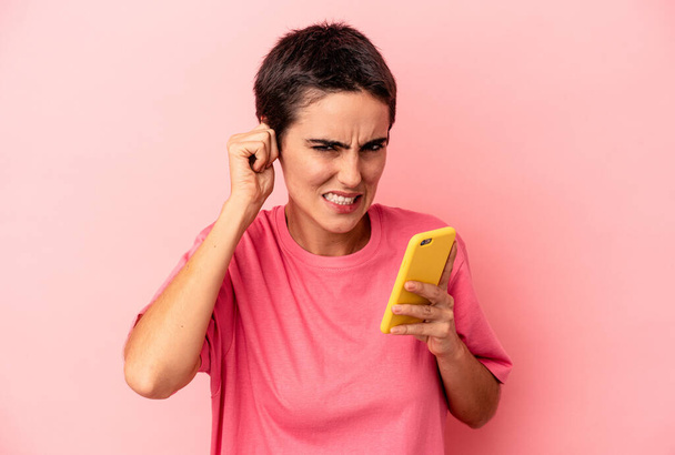 Mladá běloška drží mobilní telefon izolované na růžovém pozadí zakrývající uši s rukama. - Fotografie, Obrázek