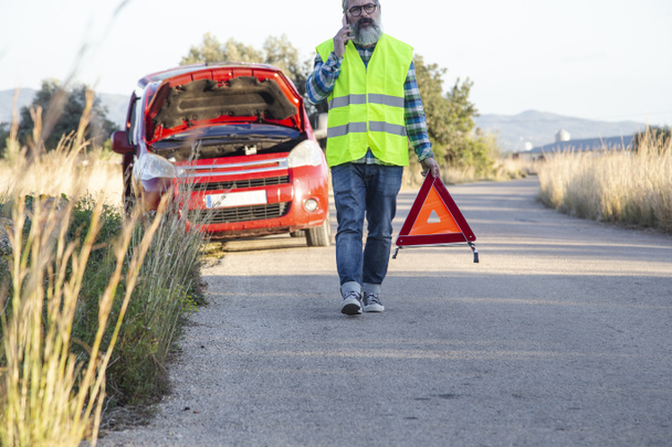 Un hombre habla por teléfono mientras pone un triángulo de advertencia detrás de su vehículo. - Foto, imagen