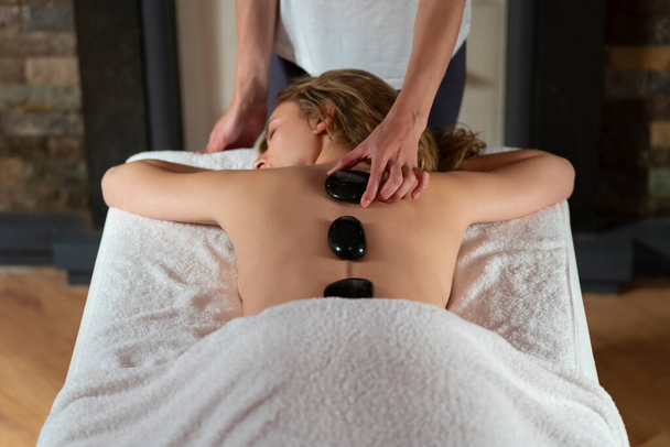 Terapeuta de massagem aplicando pedras quentes nas costas de uma mulher loira - Foto, Imagem