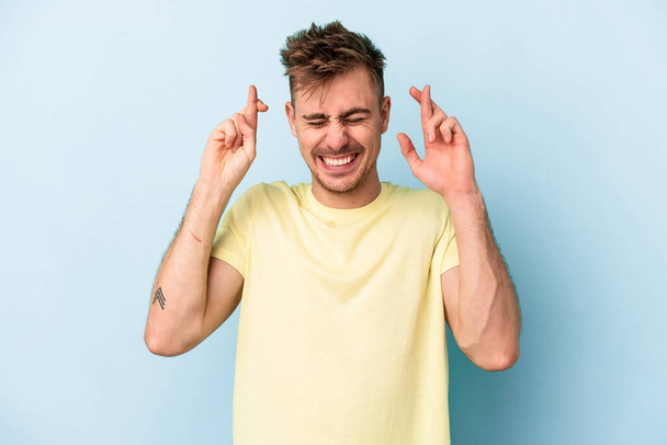 Young caucasian man isolated on blue background crossing fingers for having luck - Photo, image
