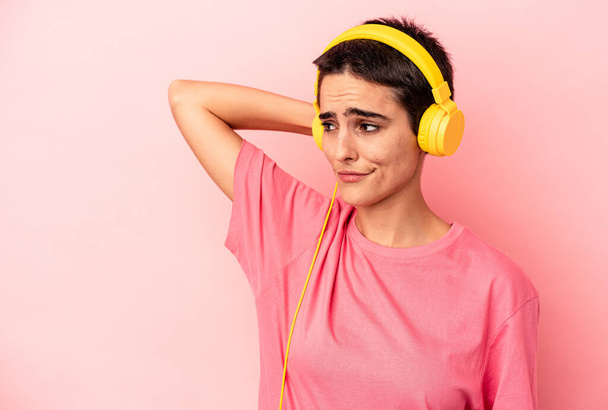 Junge kaukasische Frau, die isoliert Musik auf rosa Hintergrund hört, berührt den Hinterkopf, denkt nach und trifft eine Entscheidung. - Foto, Bild
