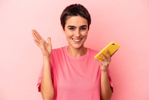 Joven mujer caucásica sosteniendo teléfono móvil aislado sobre fondo rosa recibiendo una agradable sorpresa, excitada y levantando las manos. - Foto, Imagen