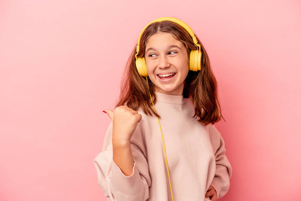Klein blank meisje luisteren naar muziek geïsoleerd op roze achtergrond punten met de duim vinger weg, lachen en zorgeloos. - Foto, afbeelding