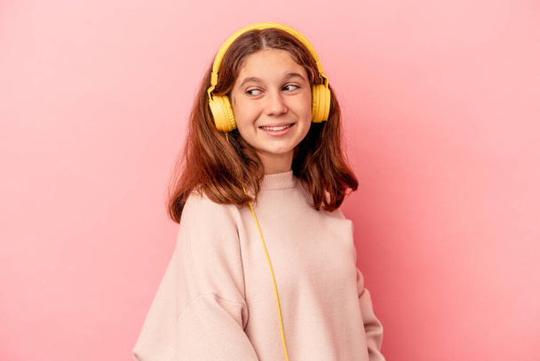 Little caucasian girl listening to music isolated on pink background looks aside smiling, cheerful and pleasant. - Photo, Image