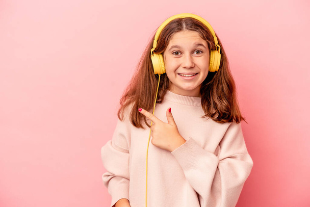 Klein blank meisje luisteren naar muziek geïsoleerd op roze achtergrond glimlachen en wijzen opzij, tonen iets op lege ruimte. - Foto, afbeelding