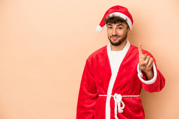 Young arab man disguised as santa claus isolated on beige background showing number one with finger. - Photo, Image