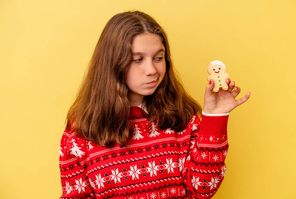 Sarı arka planda izole edilmiş bir Noel kurabiyesi tutan küçük beyaz kız kafası karışmış ve kendinden emin değil.. - Fotoğraf, Görsel