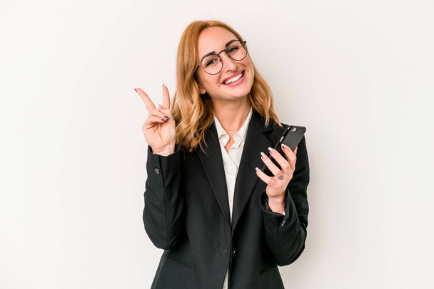 Young business caucasian woman holding mobile phone isolated on white background joyful and carefree showing a peace symbol with fingers. - Photo, image