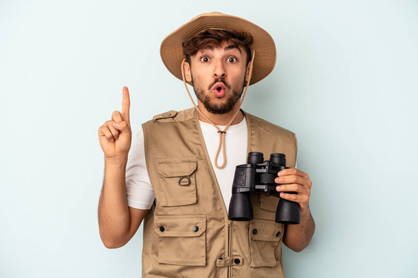 Young mixed race man holding binoculars isolated on blue background having some great idea, concept of creativity. - 写真・画像