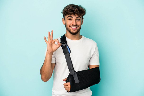 Young arab man with broken hand isolated on blue background cheerful and confident showing ok gesture. - Fotoğraf, Görsel