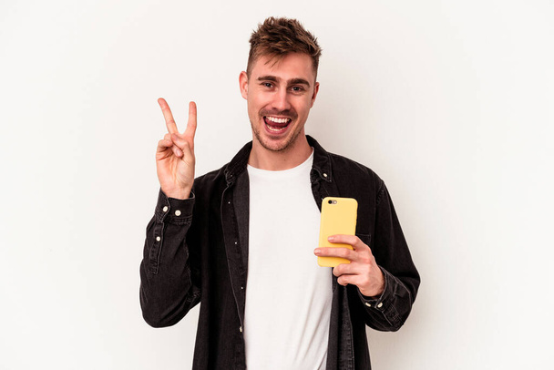 Jovem caucasiano segurando um telefone celular isolado em fundo branco alegre e despreocupado mostrando um símbolo de paz com os dedos. - Foto, Imagem