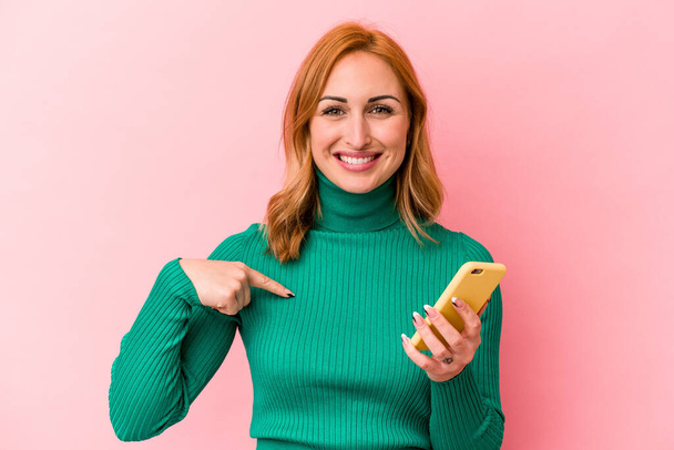 Young caucasian woman holding a mobile phone isolated on pink background person pointing by hand to a shirt copy space, proud and confident - Fotó, kép
