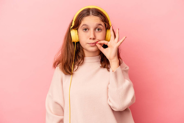 Pequeña chica caucásica escuchando música aislada sobre fondo rosa con los dedos en los labios manteniendo un secreto. - Foto, imagen