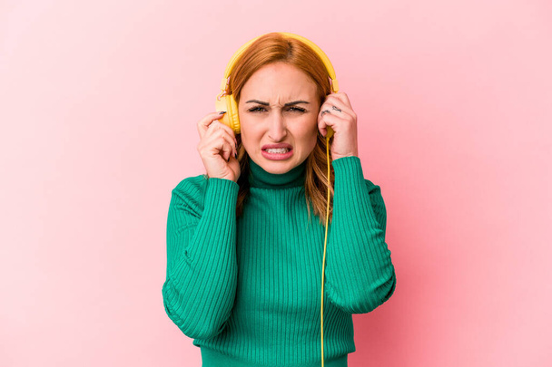 Junge kaukasische Frau hört isoliert Musik auf rosa Hintergrund, die Ohren mit Händen bedeckt. - Foto, Bild