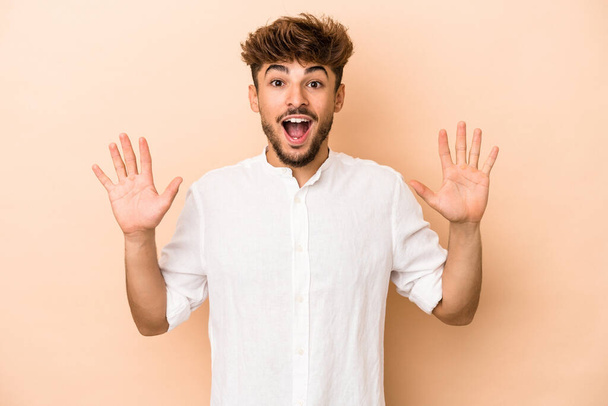Young arab man isolated on beige background receiving a pleasant surprise, excited and raising hands. - Φωτογραφία, εικόνα