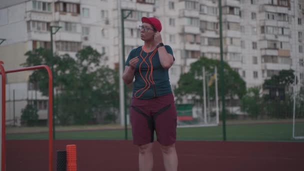 Sports theme, music in headphones, overweight cardio workout and weight loss goal. Overweight woman puts on headphones and turns on music before training jumping rope at city stadium in summer - Footage, Video