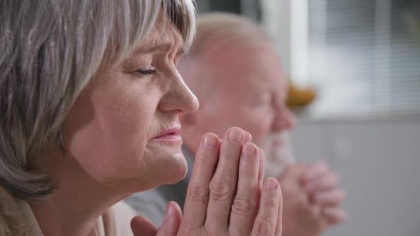 geloof, een oudere vrouw met haar oude man bidden met gesloten ogen en gevouwen handen, staande op het rijtuig met beven en tederheid voor God in de kamer, close-up - Video