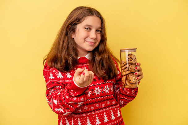 Little caucasian girl holding a Christmas cookies isolated on yellow background pointing with finger at you as if inviting come closer. - Photo, Image