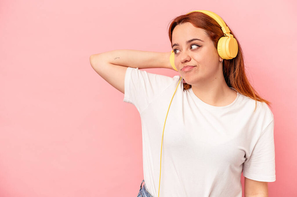 Jovem caucasiana ouvindo música isolada no fundo rosa tocando parte de trás da cabeça, pensando e fazendo uma escolha. - Foto, Imagem