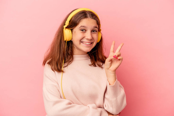 Petite fille caucasienne écoutant de la musique isolée sur fond rose montrant le numéro deux avec les doigts. - Photo, image