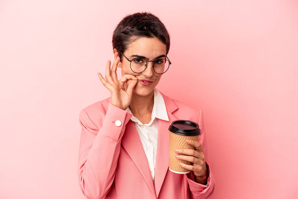Jonge blanke zakenvrouw houdt een afhaalkoffie geïsoleerd op roze achtergrond met vingers op de lippen het houden van een geheim. - Foto, afbeelding