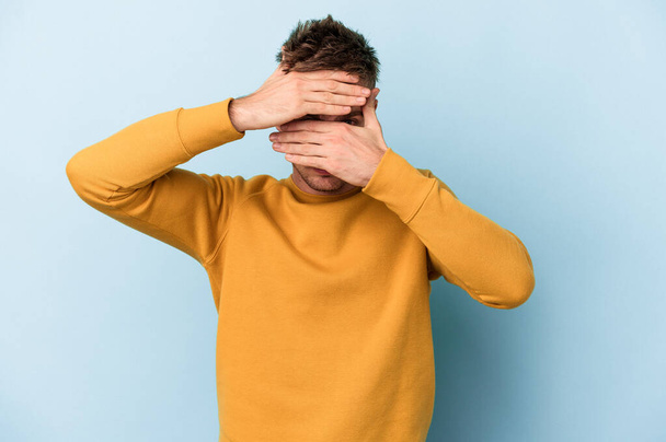 Young caucasian man isolated on blue background blink at the camera through fingers, embarrassed covering face. - Foto, Imagem