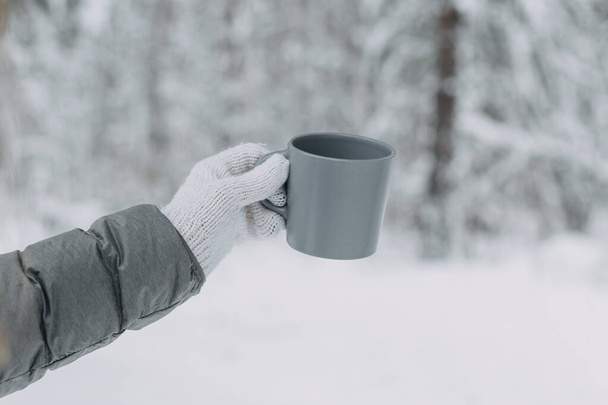 A hand in a white knitted woolen mitt and a gray jacket holds a gray mug against the background of a beautiful snow-covered forest.selective focus. - Foto, Imagem