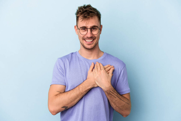 Young caucasian man isolated on blue background laughing keeping hands on heart, concept of happiness. - Photo, Image