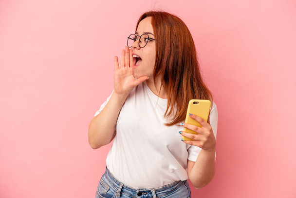 Junge kaukasische Frau hält ein Handy isoliert auf rosa Hintergrund schreit und hält Handfläche in der Nähe geöffneten Mundes. - Foto, Bild