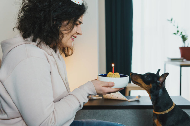 Happy young girl giving homemade cake to her dog, indoors. Concept of celebrating dog's birthday party - Foto, immagini
