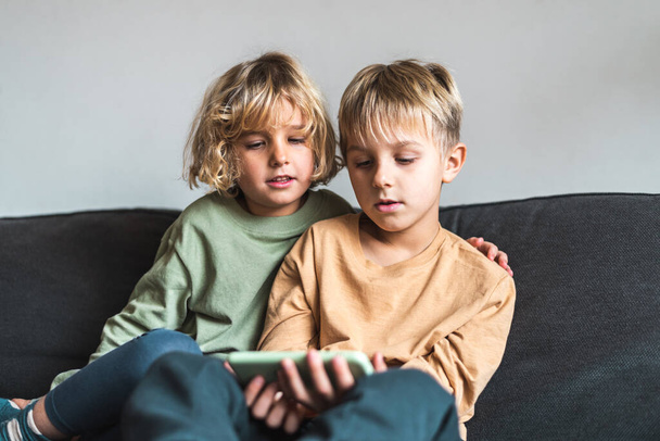 Cute little kids sharing smartphone on cozy sofa at home - Photo, Image