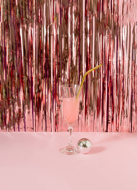 A glass of  pink drink with disco ball against shiny rose gold tassles. - Foto, afbeelding