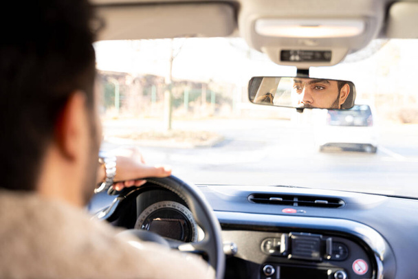 Άντρας που κρατά το τιμόνι οδηγώντας αυτοκίνητο κοιτάζοντας στον καθρέφτη οπισθοπορείας την ημέρα - Φωτογραφία, εικόνα
