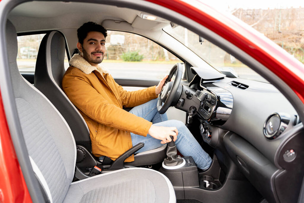 Eastern man in jacket holding driving wheel and manual gear box riding car - Foto, Imagen